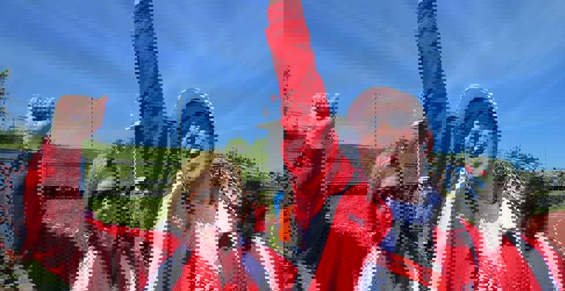Two people in red jackets are cheering happily for the camera with one arm each, raised up. One individual is an athlete as he is also wearing the number 322 pinned to his shirt and holding a blue water bottle.
