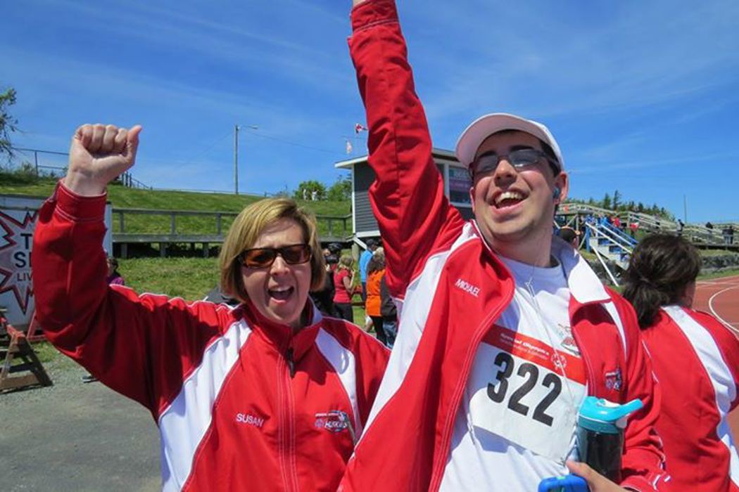 Two people in red jackets are cheering happily for the camera with one arm each, raised up. One individual is an athlete as he is also wearing the number 322 pinned to his shirt and holding a blue water bottle.