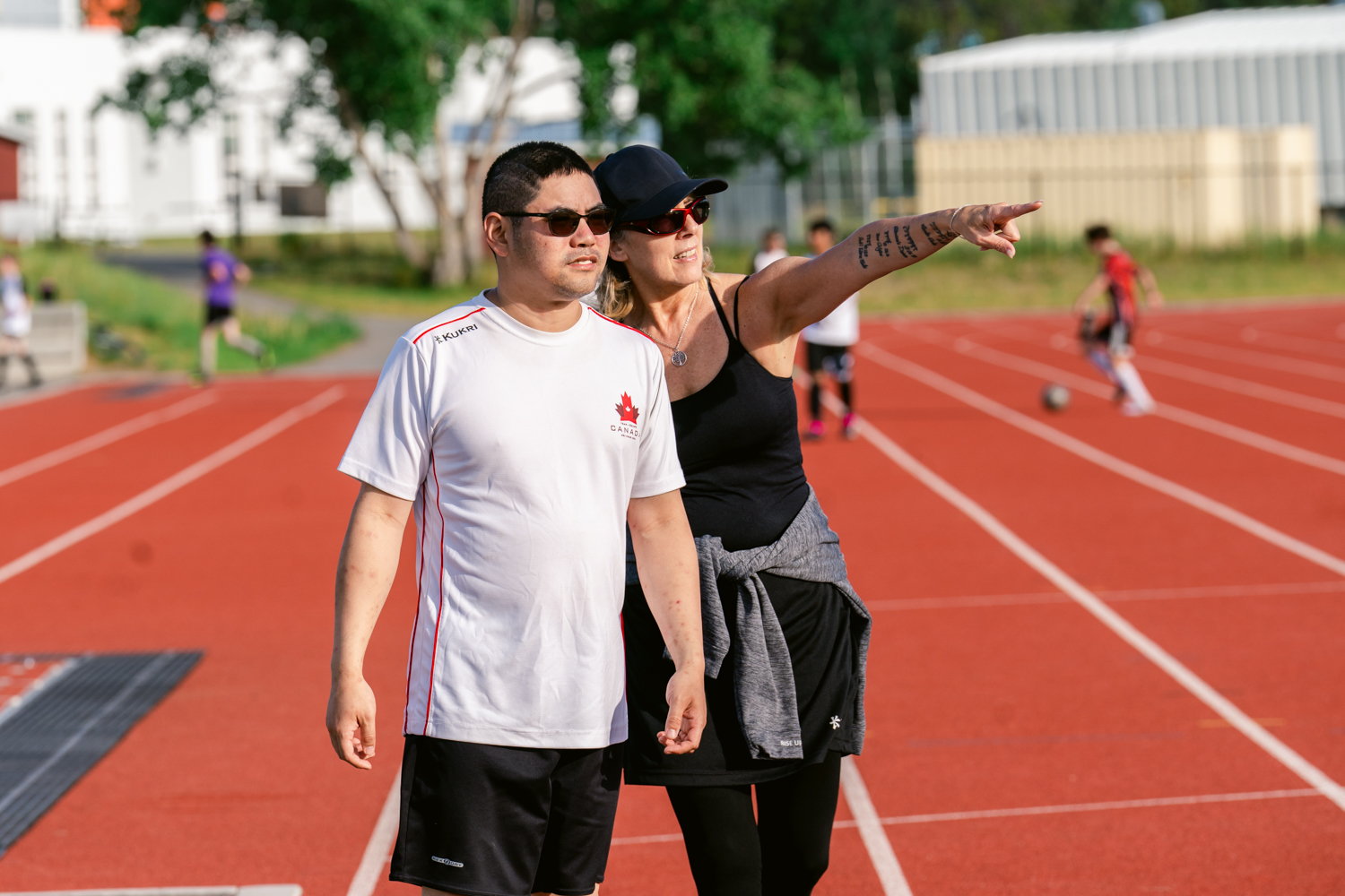 A coach is standing behind an athlete and pointing to the side, giving direction to the athlete. They are standing on a track. The coach is wearing all black with a black baseball cap. The athlete is wearing a white t-shirt and sunglasses.
