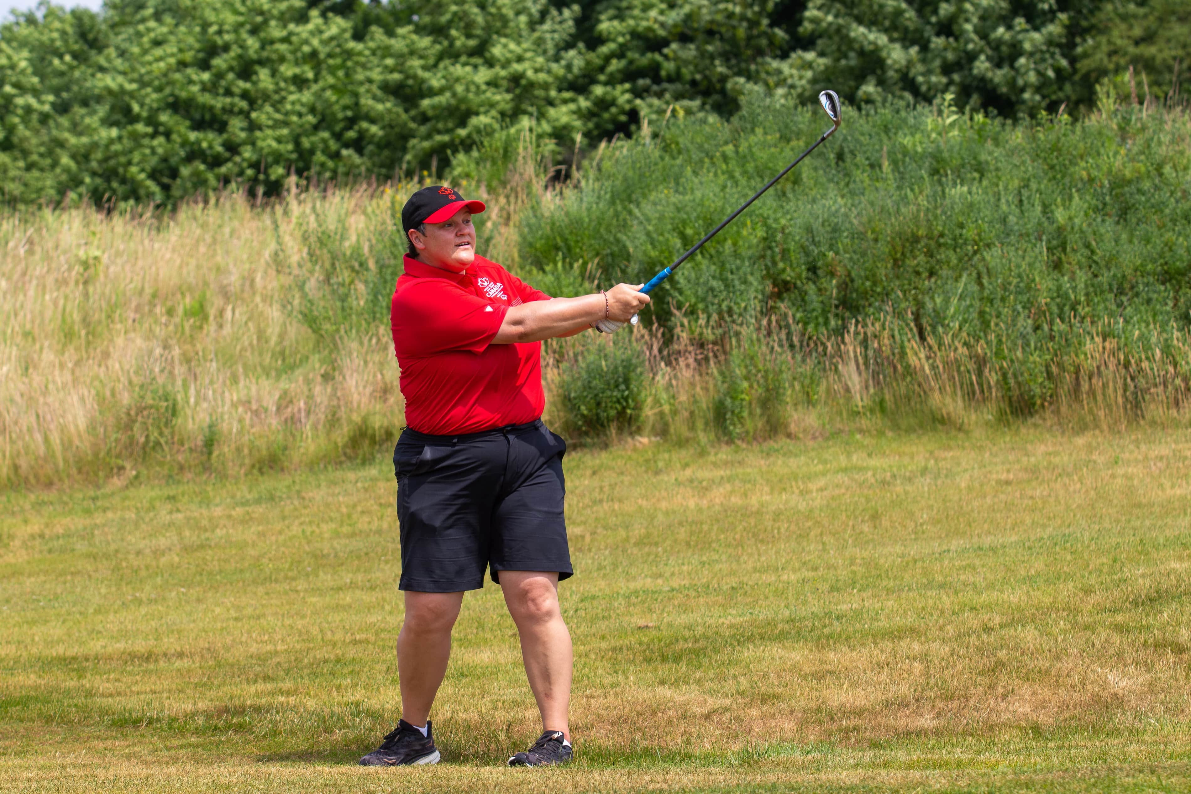 Person following through on a golf swing, dressed in a red golf shirt and hat and black shorts. Person following through on a golf swing, dressed in a red golf shirt and hat and black shorts.