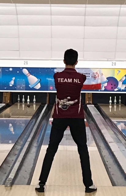 A bowling athlete is standing at the top of the bowling aisle, preparing for their turn. The camera is directly behind the athlete. The athlete is wearing a burgundy 