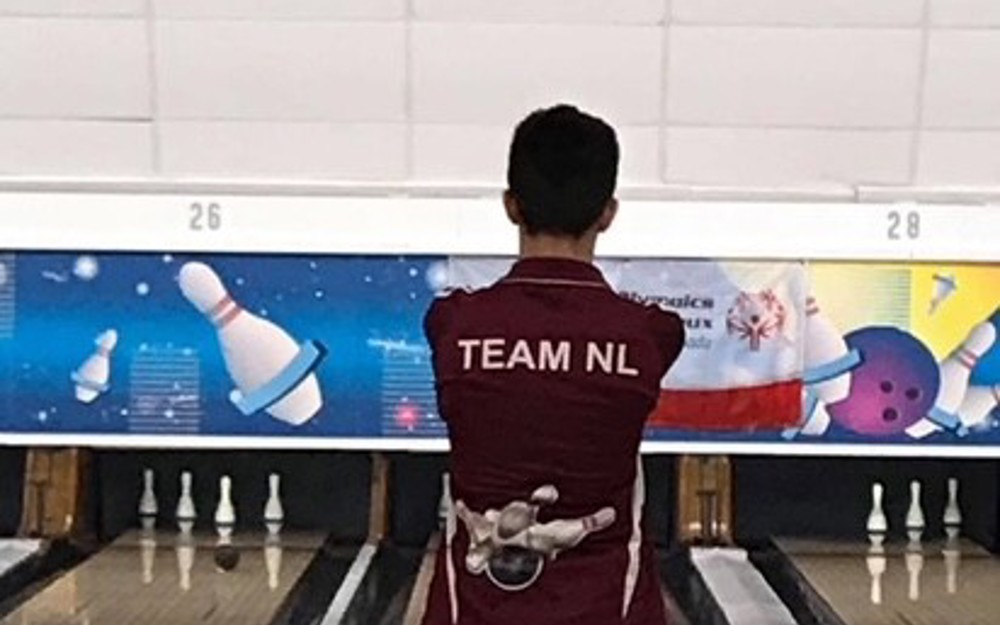 A bowling athlete is standing at the top of the bowling aisle, preparing for their turn. The camera is directly behind the athlete. The athlete is wearing a burgundy "Team NL" bowling shirt.