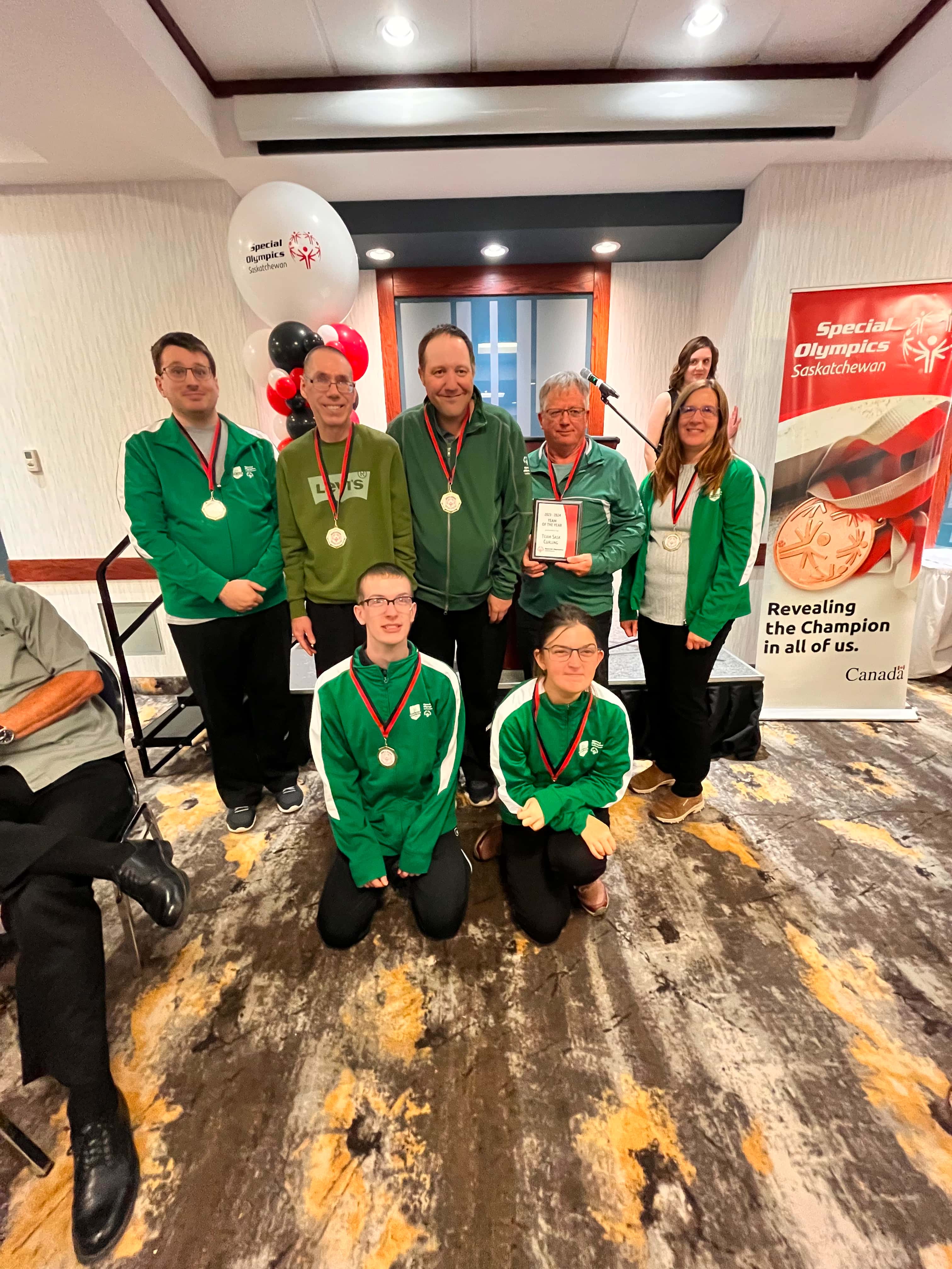 A group team photo of athletes who have won medals. They pose, two people in front on their knees and five standing behind them. They are all wearing green sweaters with a ribboned medal hanging around their necks. The image takes place in a large room with banners and balloons behind the team.