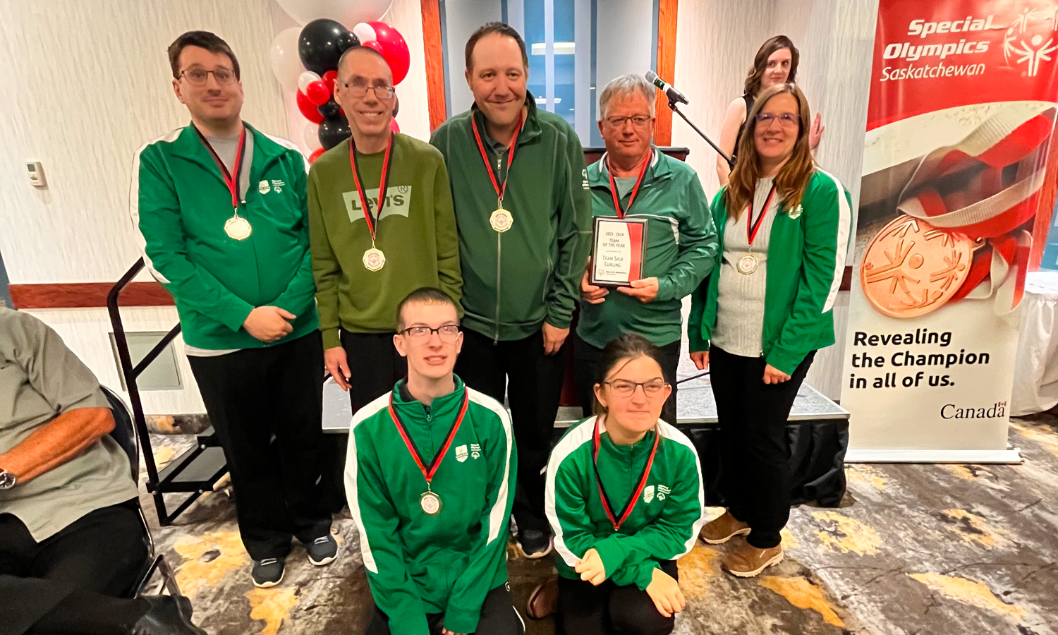 A group team photo of athletes who have won medals. They pose, two people in front on their knees and five standing behind them. They are all wearing green sweaters with a ribboned medal hanging around their necks. The image takes place in a large room with banners and balloons behind the team.