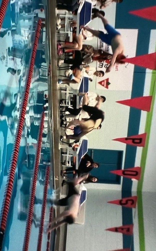 Three Special Olympics Newfoundland & Labrador athletes are diving into an indoor swimming pool from the diving blocks at the edge of the pool. The three athletes are blurry due to being in motion.