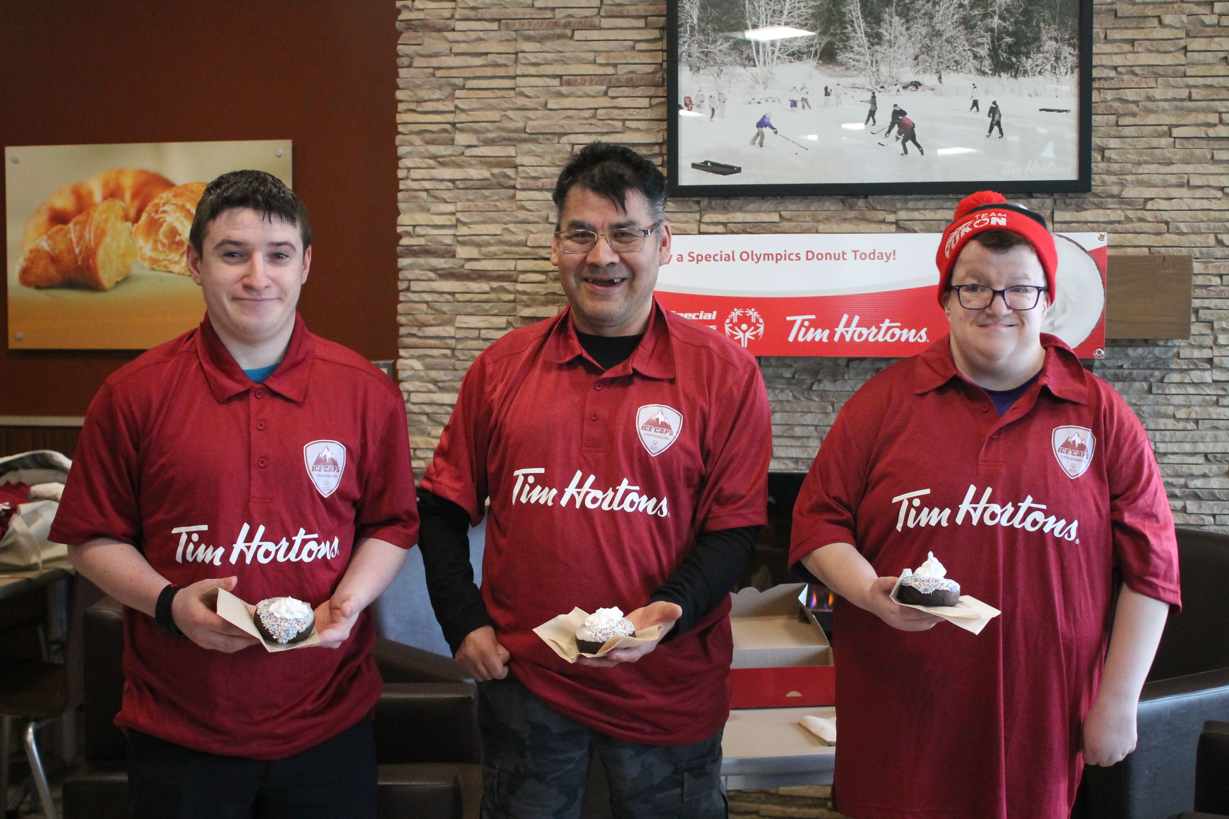 Three athletes smile and pose for the camera holding a Special Olympics donut from Tim Hortons. They are each wearing red Tim Hortons shirts and they are standing inside a Tim Hortons shop.
