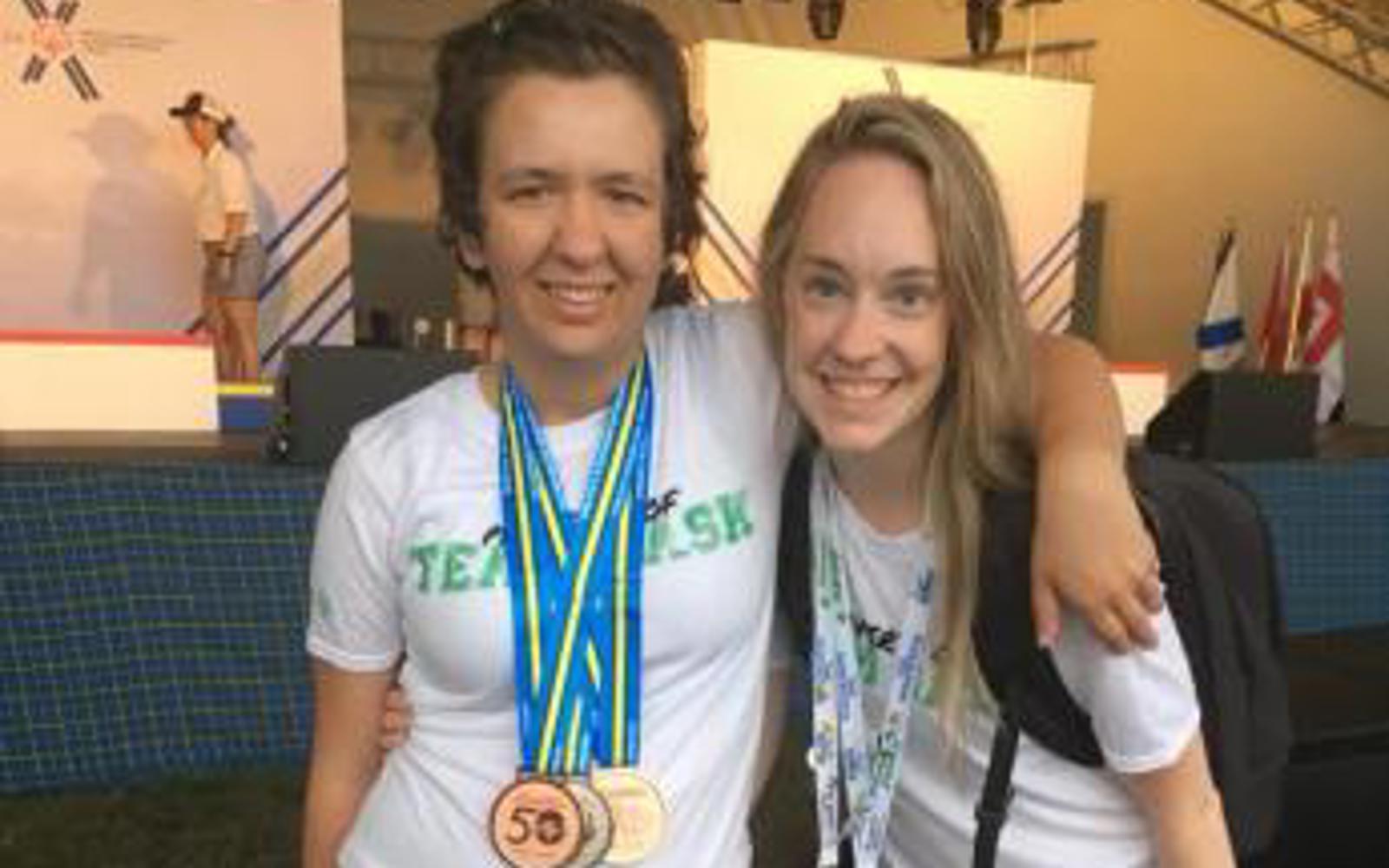 A photo of a volunteer and athlete with their arms around behind each other smiling for the camera. They are both wearing white t-shirts. The athlete has medals around her neck while the volunteer has a colourful lanyard around her neck.