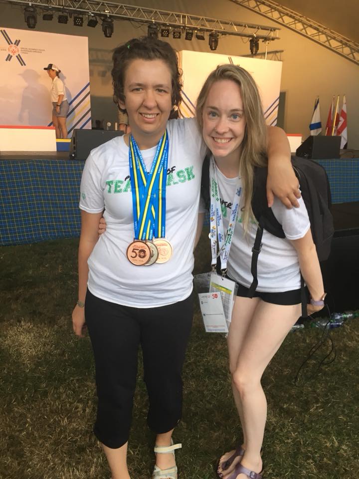 A photo of a volunteer and athlete with their arms around behind each other smiling for the camera. They are both wearing white t-shirts. The athlete has medals around her neck while the volunteer has a colourful lanyard around her neck. A photo of a volunteer and athlete with their arms around behind each other smiling for the camera. They are both wearing white t-shirts. The athlete has medals around her neck while the volunteer has a colourful lanyard around her neck.