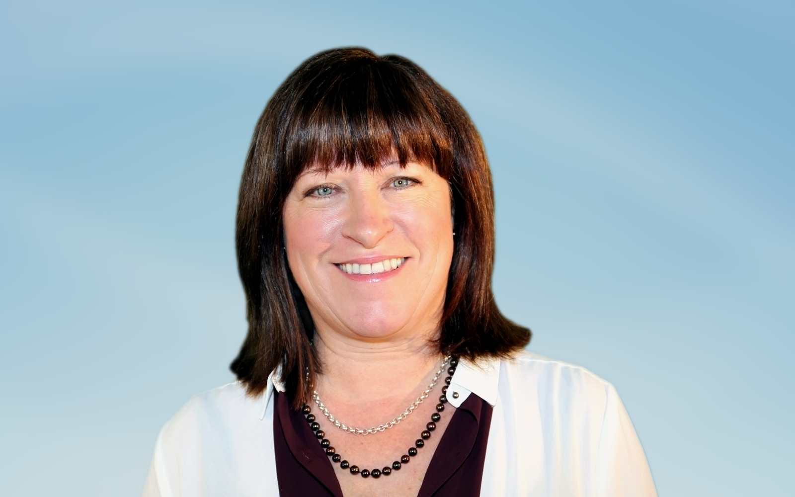 Femme vêtue d'un chemisier noir et d'un blazer blanc. Elle porte deux colliers : un collier de perles noires et une chaîne en argent. Elle sourit à la caméra.