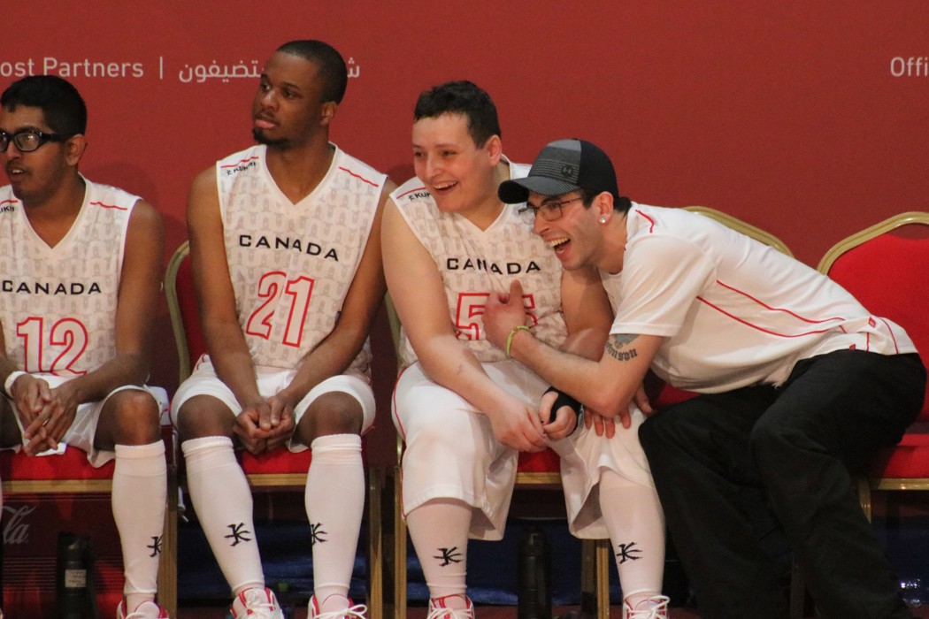 Quatre hommes sont assis sur un banc. Ils sont tous vêtus d'uniformes de basketball de l'équipe canadienne blancs et rouges. L'un des hommes est penché en avant et encourage les joueurs. 