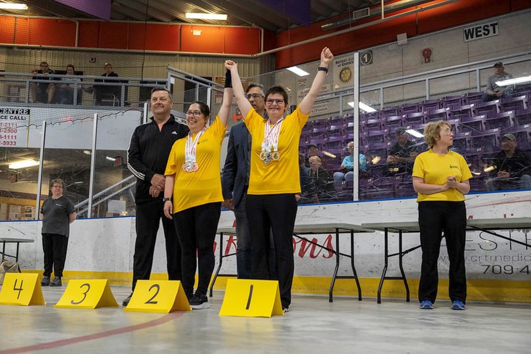 Two winning athletes are standing next to each other holding hands, with their arms raised in the air. They are both wearing yellow t-shirts and have multiple medals hanging around their necks. There are paper signs in front of them that read "1", "2", "3", "4" from right to left.