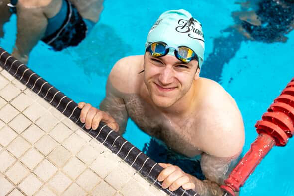 A bird eye view of Maxim Thibault smiling up at the camera. He is in a swmming pool wearing a swim cap on his head and goggles on his forehead. 