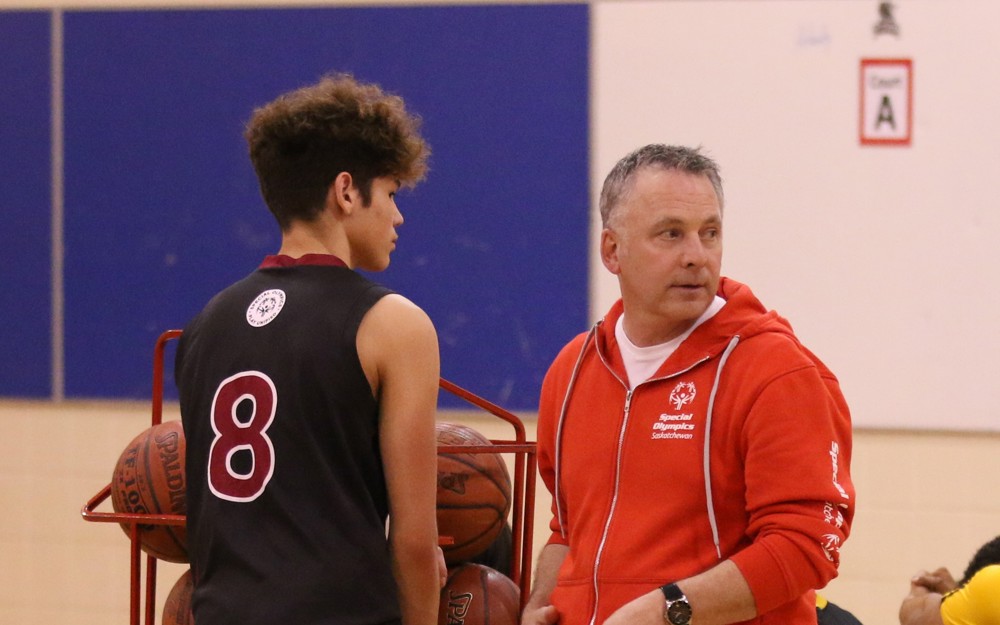 A basketball coach talking to a basketball athlete. They are standing to the side of the court next to a rack filled with basketballs.