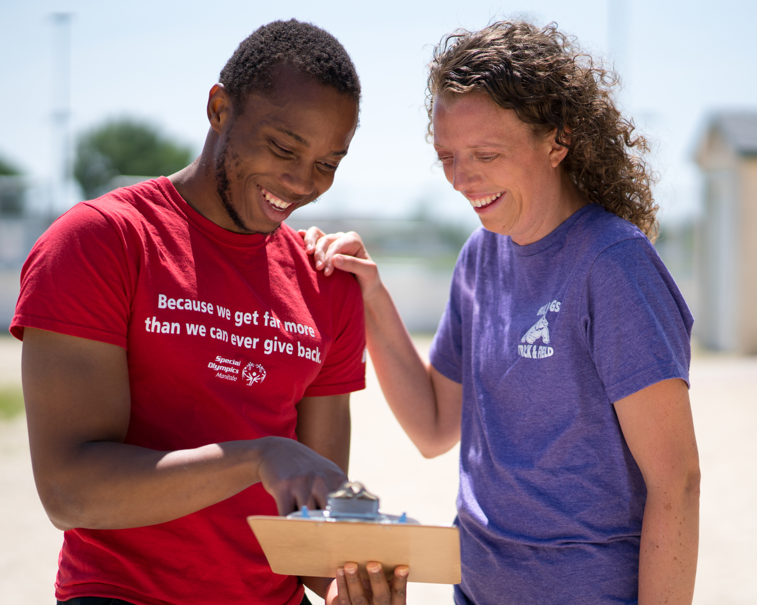 A person in a red shirt with the words "Because we get far more than we can ever give back" holding and pointing at a clipboard while a person in a purple shirt smiles down at the clipboard. 