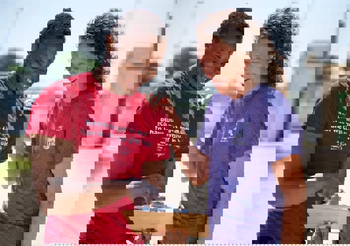 Une personne portant une chemise rouge avec les mots « Parce que nous recevons bien plus que nous ne pourrons jamais donner en retour » tient et montre un bloc-notes tandis qu'une personne portant une chemise violette sourit en regardant le bloc-notes. 