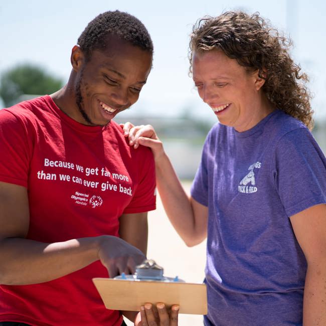 A person in a red shirt with the words "Because we get far more than we can ever give back" holding and pointing at a clipboard while a person in a purple shirt smiles down at the clipboard. 
