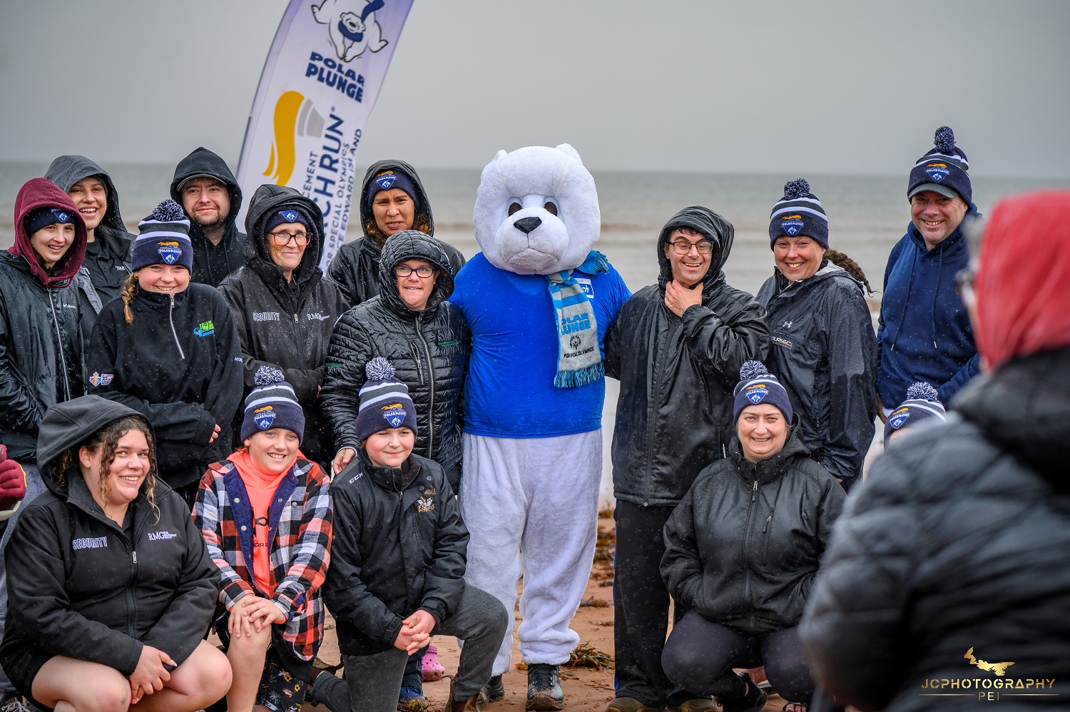 A group of people stand on a beach with a mascot after participating in a Polar Plunge