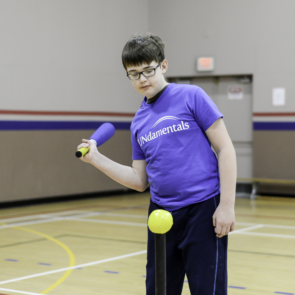 A young athlete in a blue FUNdamentals t-shirt, is swinging back a baseball bat to hit a yellow ball perched on a post. He is in a gymnasium and wearing a pair of glasses.