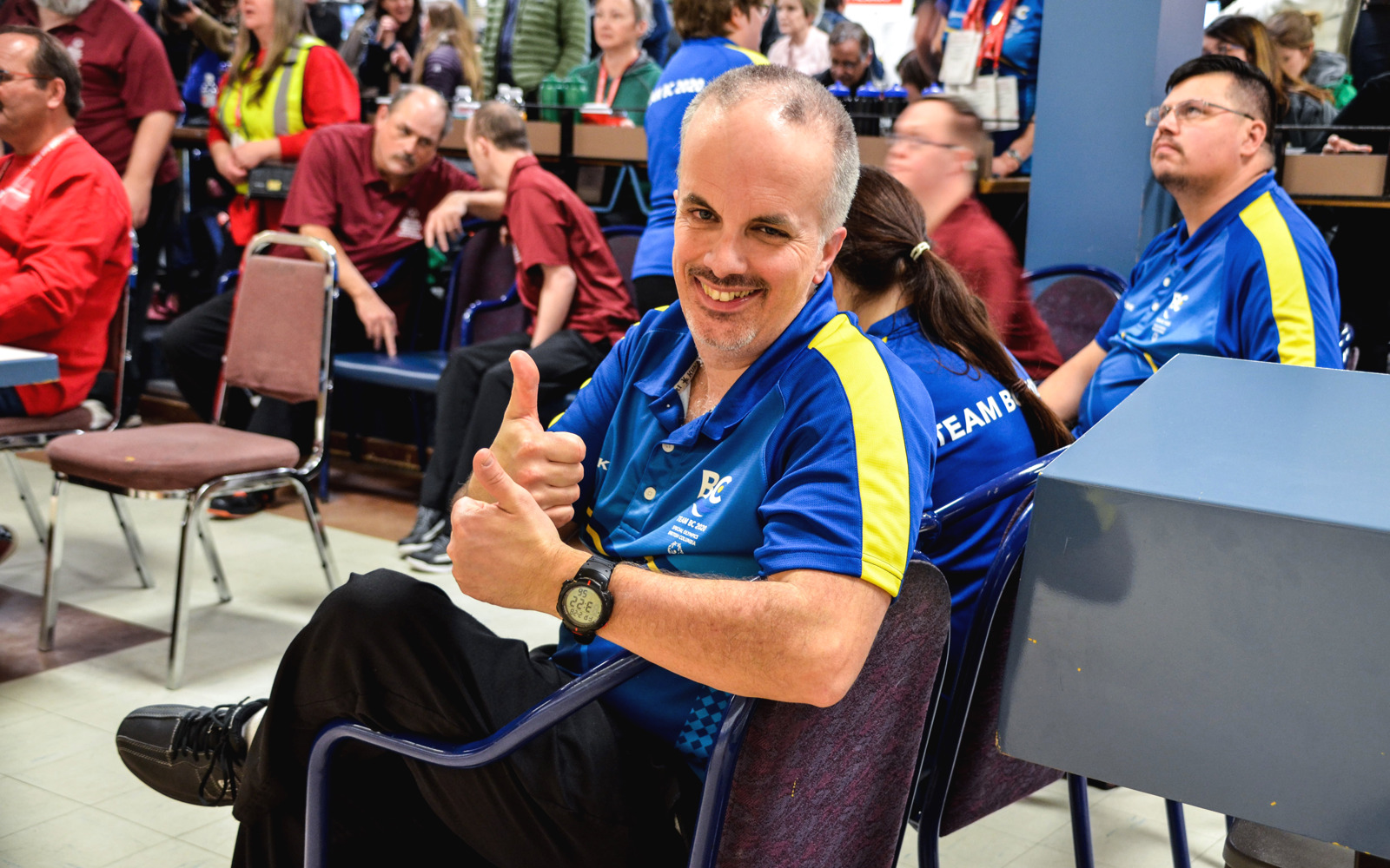 A Special Olympics BC bowling athlete takes a seat on a chair and turns to the camera with a smile and a thumbs up.