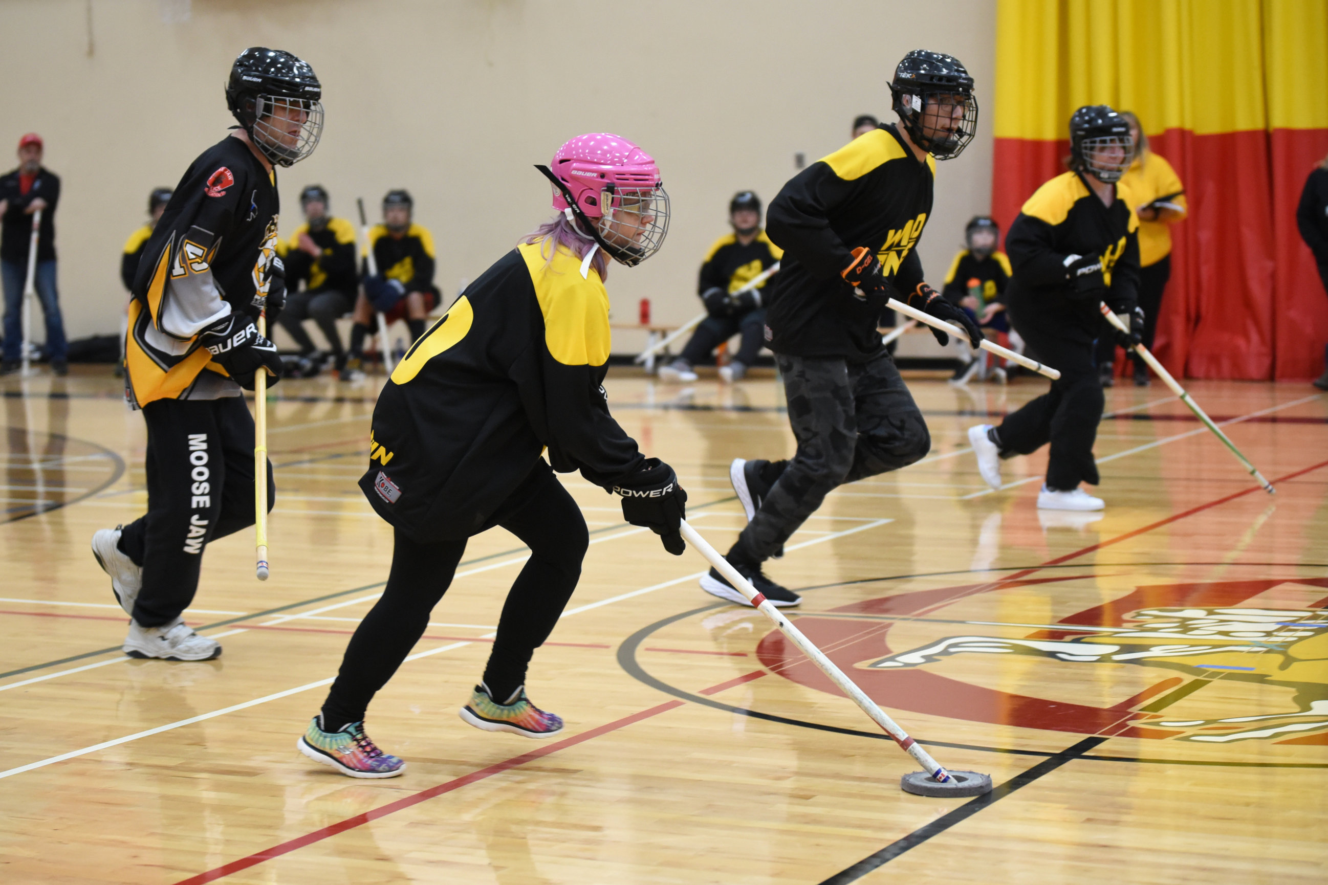 An action shot photo of athletes playing floor hockey. Four athletes can be seen running across the gym floor with their floor hockey sticks. They are all wearing helmets and black and yellow floor hockey jerseys.