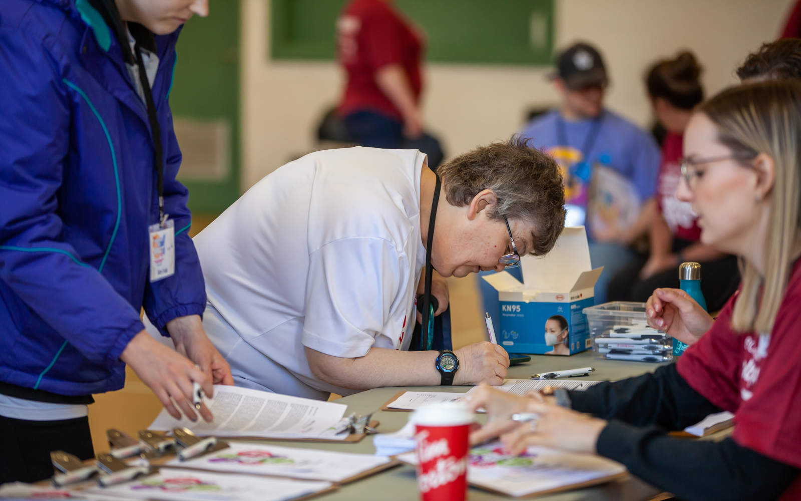 Two athletes are at a volunteer table filling out forms. One athlete, in the camera's focus, is leaning over a table filling out forms. The other athlete is standing up reviewing the forms. A volunteer sits at the table assisting the athletes in filling out their forms.