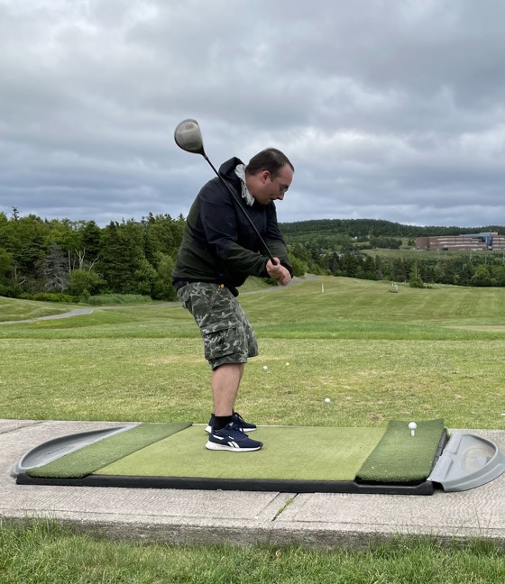 An action shot photo of a golf athlete taking a swing at the start of the golf game. It is a cloudy day and the athlete is wearing a black sweater. In the distance you can see the other golf balls that have been in play.
