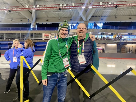 An athlete posing with his coach after the game. They are both smiling for the camera with their arms wrapped behind each other. The athlete is wearing a green sweater, a pair of jeans, and a winter hat. The coach is wearing a navy vest, and a teal sweater. They both have a lanyard around their necks.