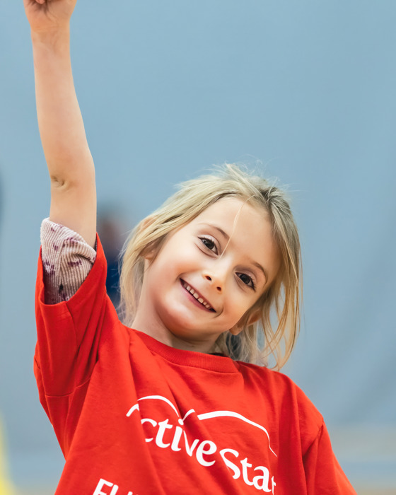 Young blond girl wearing a red shirt with her arm in the air smiling at the camera. 