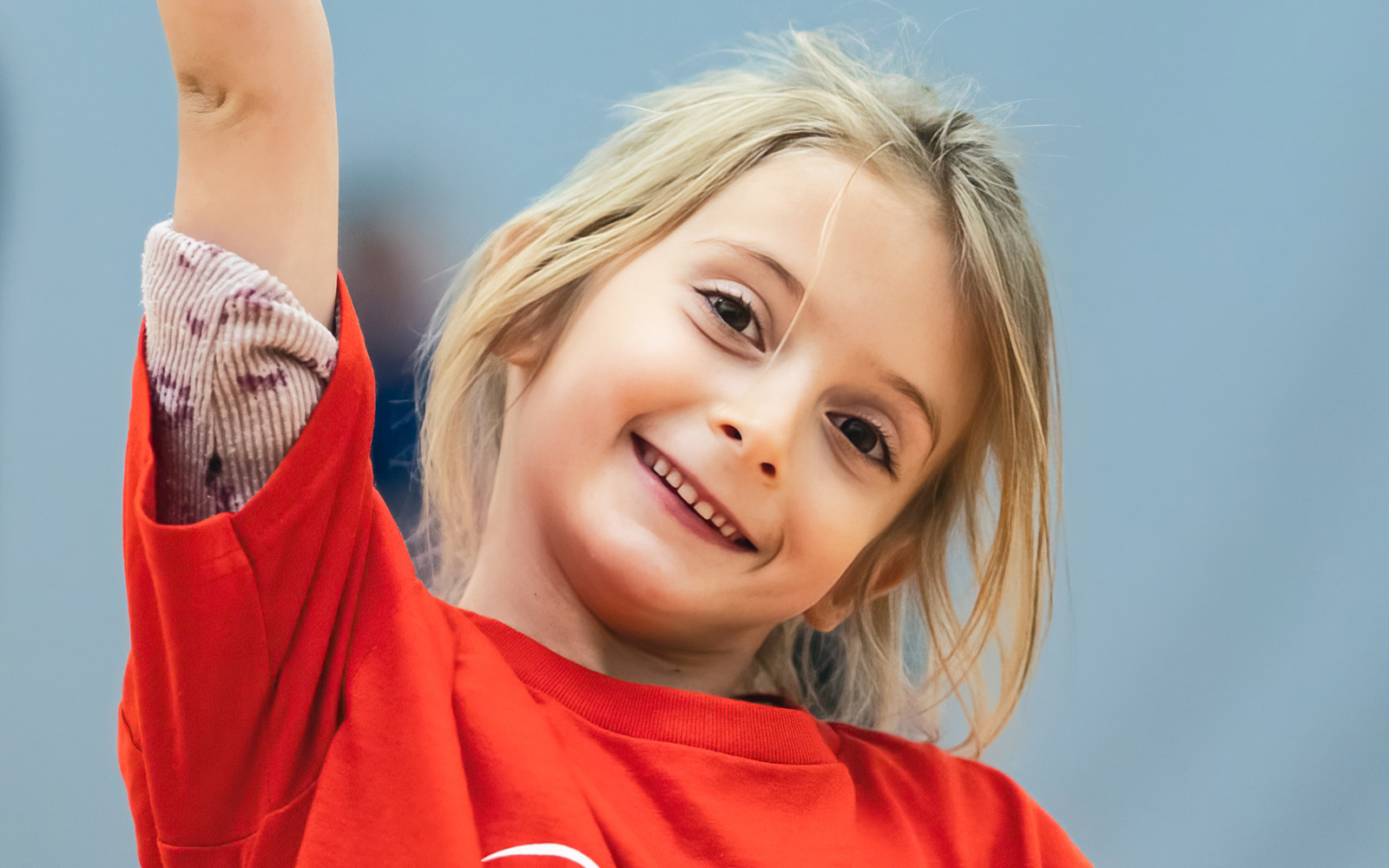 Young blond girl wearing a red shirt with her arm in the air smiling at the camera.