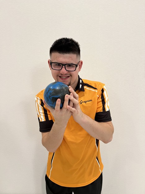 Young man in a yellow bowling shirt with glasses is holding a blue bowling ball like he is about to throw it and smiling at the camera. 