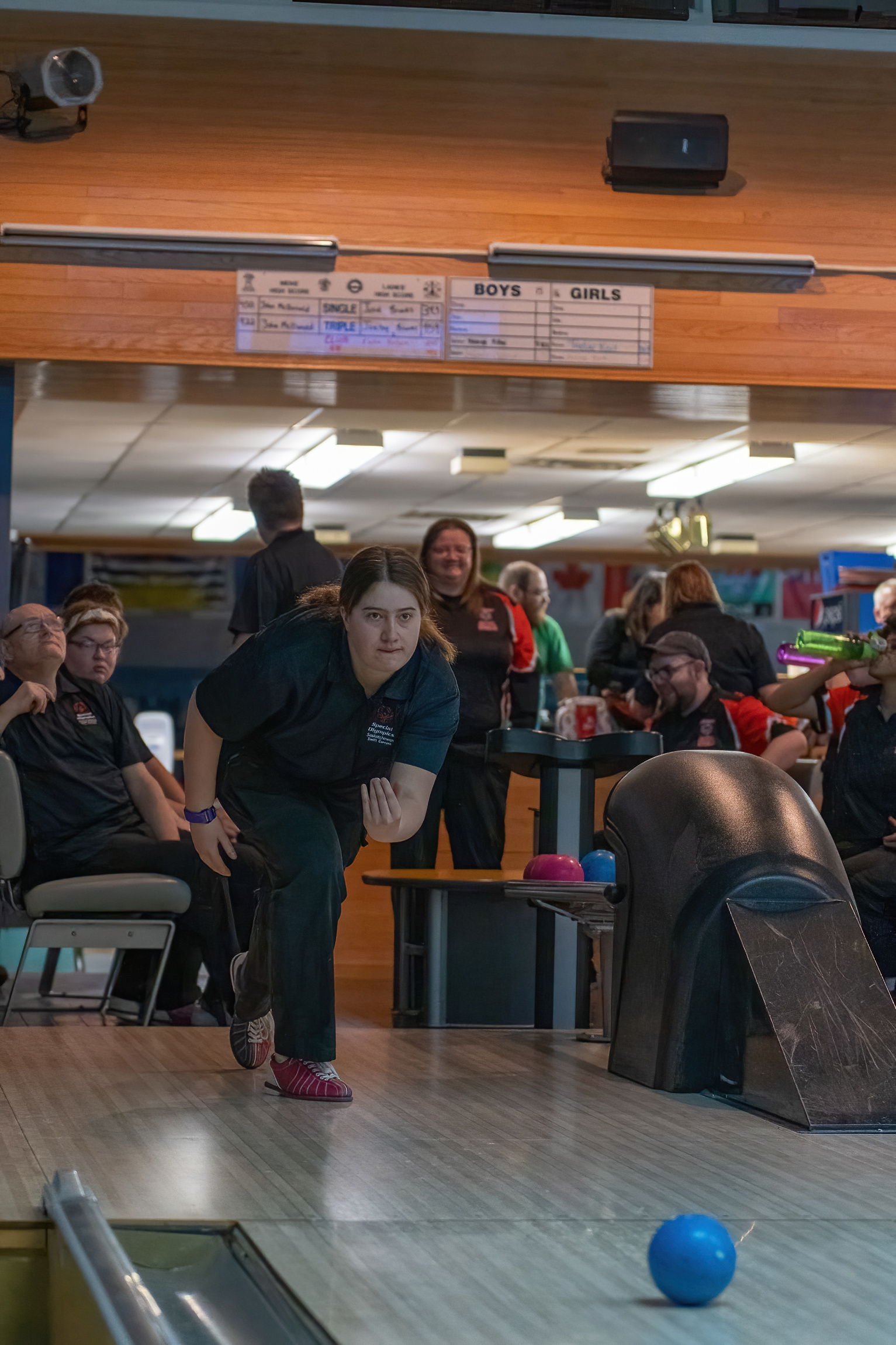 An action shot photo of a bowling athlete. She is at the start of the lane, mid throw, and you can see the blue bowling ball start to roll down the aisle. Behind the athlete, you can see her teammates smiling and watching her turn. An action shot photo of a bowling athlete. She is at the start of the lane, mid throw, and you can see the blue bowling ball start to roll down the aisle. Behind the athlete, you can see her teammates smiling and watching her turn.