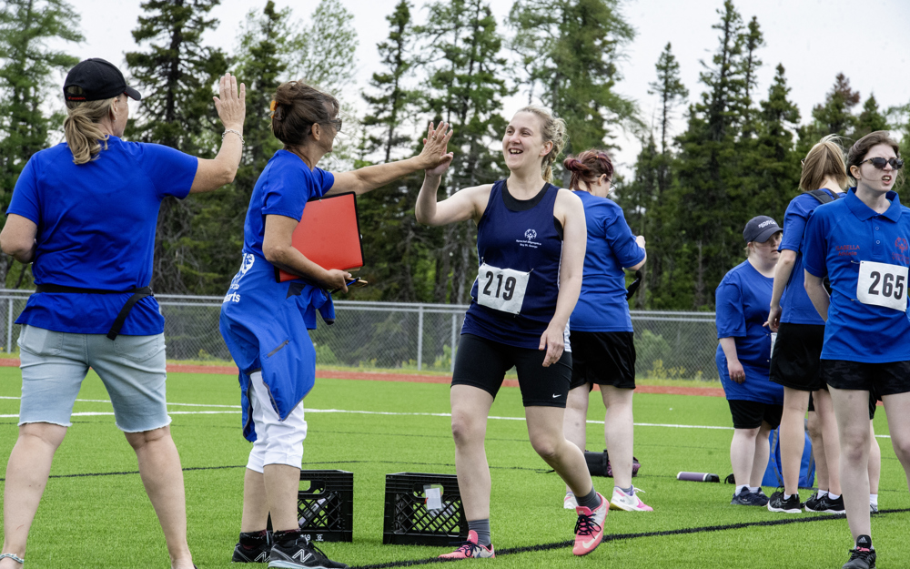 An athlete, on the sidelines after her run, is high-fiving with her coach. Her coach is holding a red binder and wearing a blue t-shirt with a red jacket wrapped around her waste. The athlete is wearing a blue tank top with black biker shorts. She has the number 219 pinned to her shirt.