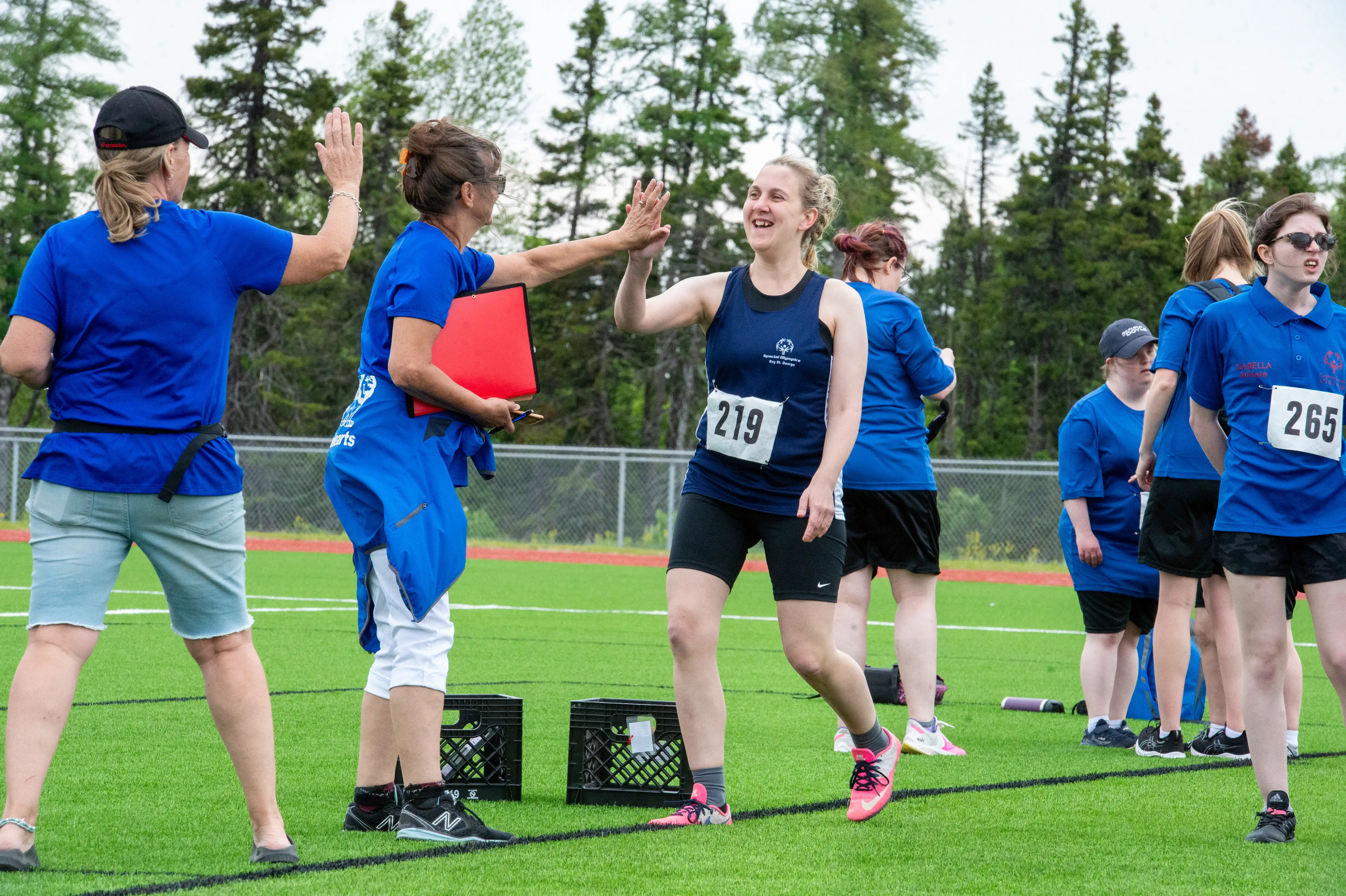 An athlete, on the sidelines after her run, is high-fiving with her coach. Her coach is holding a red binder and wearing a blue t-shirt with a red jacket wrapped around her waste. The athlete is wearing a blue tank top with black biker shorts. She has the number 219 pinned to her shirt. An athlete, on the sidelines after her run, is high-fiving with her coach. Her coach is holding a red binder and wearing a blue t-shirt with a red jacket wrapped around her waste. The athlete is wearing a blue tank top with black biker shorts. She has the number 219 pinned to her shirt.