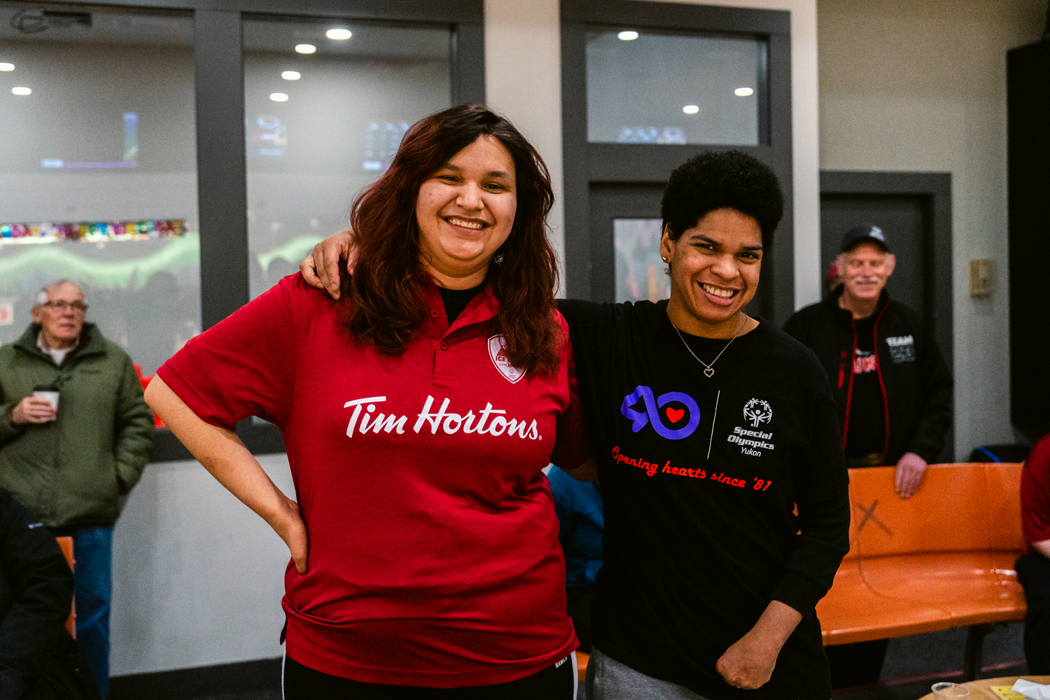 Two bowling players with their arms wrapped around behind each other, are smiling for the camera. The player on the left is wearing a red Tim Hortons collared shirt. The player on the right is wearing a black Special Olympics Yukon shirt. Two bowling players with their arms wrapped around behind each other, are smiling for the camera. The player on the left is wearing a red Tim Hortons collared shirt. The player on the right is wearing a black Special Olympics Yukon shirt.
