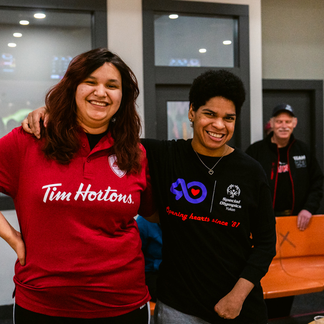 Two bowling players with their arms wrapped around behind each other, are smiling for the camera. The player on the left is wearing a red Tim Hortons collared shirt. The player on the right is wearing a black Special Olympics Yukon shirt.