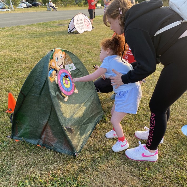 Photo taken outdoors. A little girl, being supported by her mother, is playing with a pop up toy. 