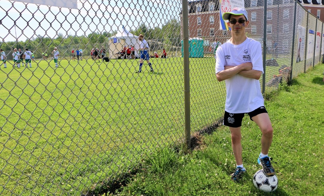 Andrew Hynes stands next to a fence with a soccer ball under his foot and arms crossed.