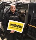 Const. Scott Edwards stands in front of a police car with his dog Chase in the front seat. Scott is holding a yellow sign that says #NoGoodWay