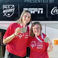 Maya and Amy lean together, holding up their medals and smiling