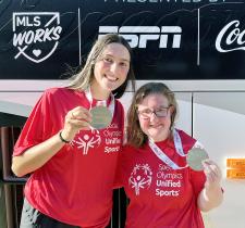 Maya and Amy lean together, holding up their medals and smiling