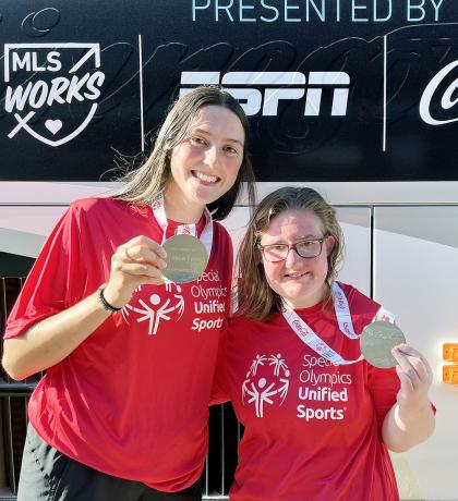 Maya and Amy lean together, holding up their medals and smiling
