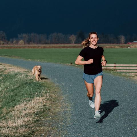 Kalyn smiling and looking strong while running down a rural path on a sunny day, followed in the distance by a running dog