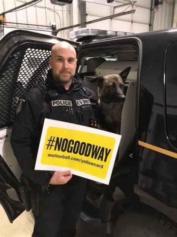 Const. Scott Edwards stands in front of a police car with the door open holding a No Good Way sign. His police dog Chase sits in the front seat.