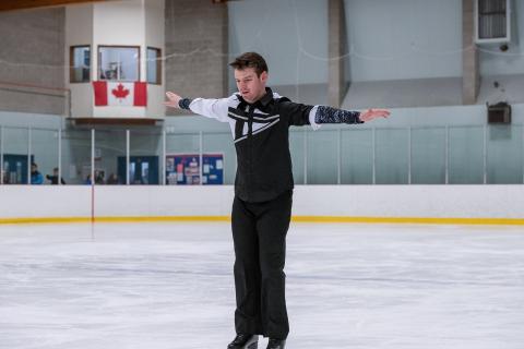 Mike competes in figure skating on the ice with his arms spread wide.