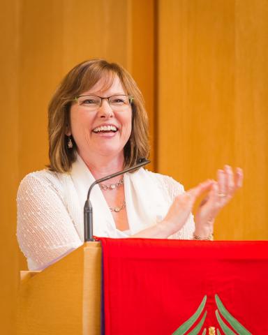 Brenda Baker speaks and claps at a podium while smiling.