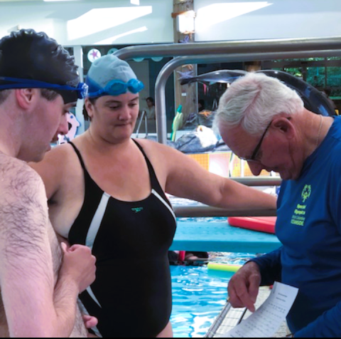 Two swimmers talking with their coach who is pointing at a clipboard for guidance